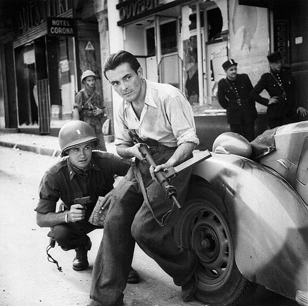 American officer and French partisan crouch behind an auto during a street fight in a French city. 1944